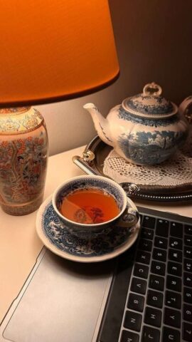 Elegant vintage tea setting with a blue-patterned tea set, lamp, and laptop on a cozy desk.