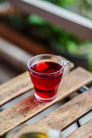 Close-up of a vibrant red herbal tea in a glass cup on a rustic wooden table outdoors.
