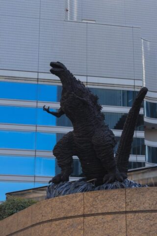 Photo by cocodillaa.com A godzilla statue stands in front of a modern building.