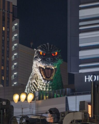 Photo by cocodillaa.com Giant godzilla statue looms over city buildings at night.