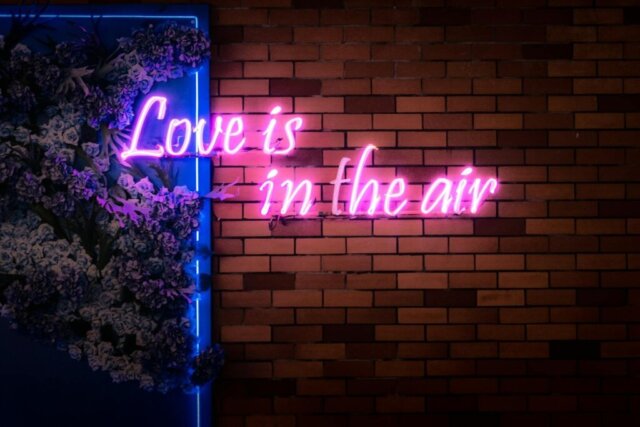 Bright neon 'Love is in the air' sign on a brick wall with floral decor in Bangkok.