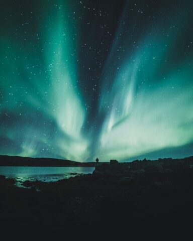 person standing near body of water during aurora northern sky person standing near body of water during aurora northern sky
