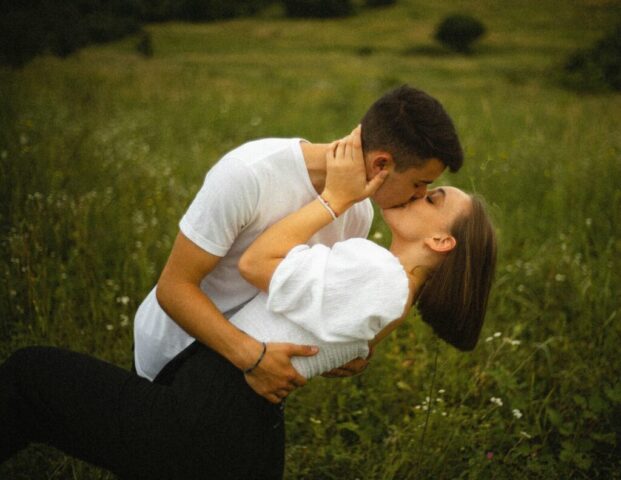 Photo by Cocodillaa.com man in white t-shirt kissing woman in white shirt on green grass field during daytime