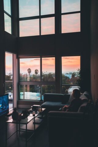Photo by Roberto Nickson woman sitting on corner sofa beside coffee table near flat screen TV inside living room with glass window walls