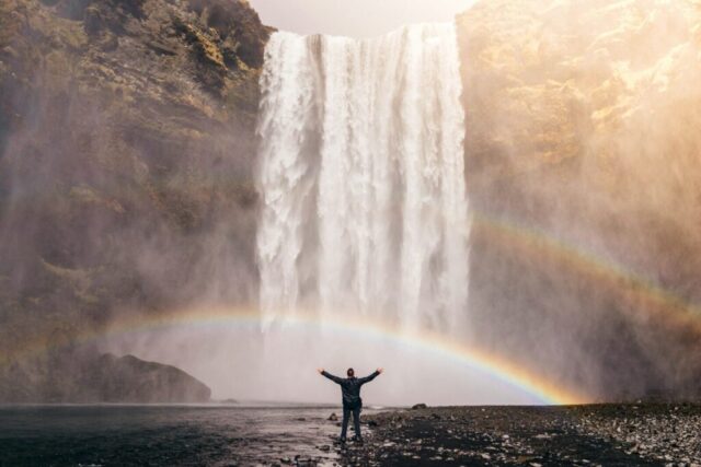 Photo by Cocodillaa.com person in front of waterfalls with double rainbow during daytime